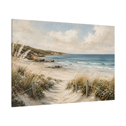 A rectangular canvas wall art print showing a coastal beach scene with a sandy path, dune grass, rocks, blue ocean water, and a cloudy sky. The canvas is photographed at a slight angle against a plain white background.