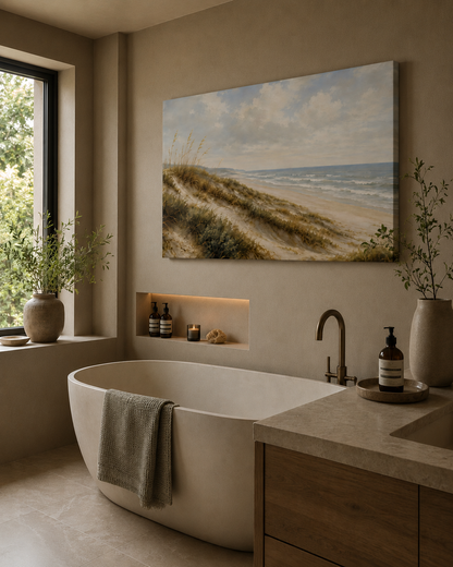 A large rectangular beach dunes landscape painting hangs on a beige wall above a freestanding white bathtub in a softly lit bathroom with a stone countertop, wood vanity, window, and plants.