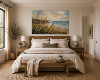 A large rectangular canvas print of a beach and dune landscape hangs above a neatly made bed in a bright, modern bedroom. The artwork shows golden beach grasses, blue ocean water, and a cloudy sky.