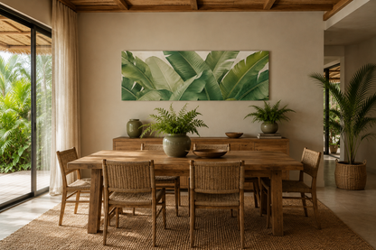 A large rectangular wall art print featuring green tropical banana leaves, displayed in a modern dining room setting above a wooden dining table and chairs.