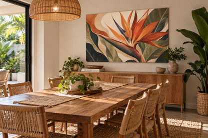 A large rectangular wall art print featuring a stylized orange bird of paradise flower with green and beige botanical leaves, displayed above a wooden dining table in a bright, modern dining room.