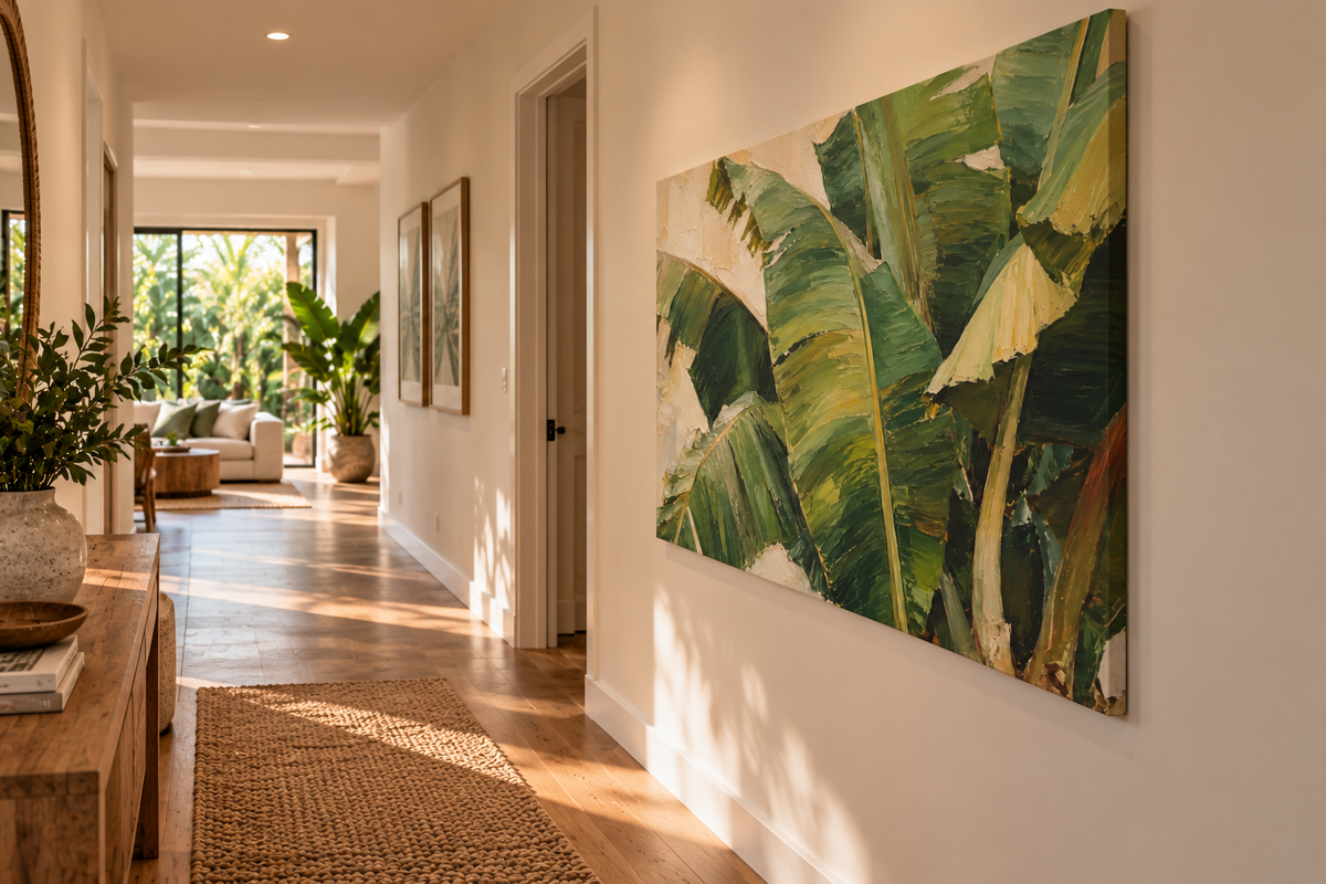 A large rectangular wall art print featuring green banana leaves hangs on a white hallway wall above a woven rug, with a sunlit interior and wood floor visible in the background.